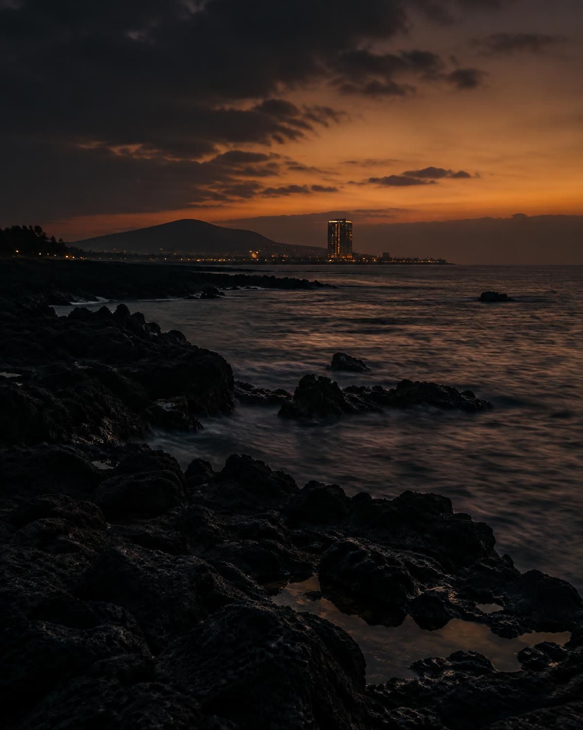 黄昏时济州海岸的黑色火山岩,远处是亮灯的酒店高楼